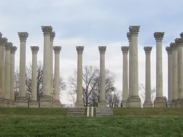 Capitol Columns standing in a wide arc on a grassy field with steps in the foreground and cloudy skies above.