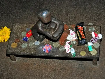 Järnpojke sculpture with a shiny head on a raised platform covered with coins, candies, chocolate bars, and a yellow flower.
