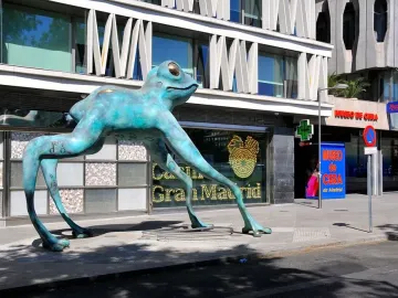 Lucky Frog sculpture with a blue-green oxidized surface and etched symbols on its legs, standing on a city sidewalk.