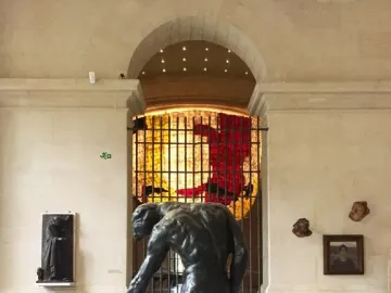 The Three Shades bronze sculpture shown in a museum gallery with a bowed male figure in the foreground.