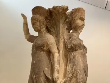 Weathered stone Dancers of Delphi with two draped female figures around a central plant-like column.