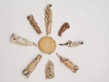 Nine carved diagnostic dolls arranged in a circle around a woven spherical object, with figures in pale ivory and darker tones.