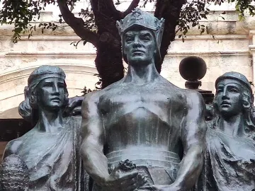 Great Siege Monument with three bronze draped figures on a white base beside a street lamp and cannon.