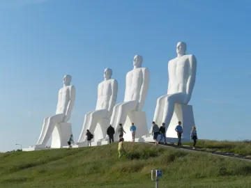 The Man by the Sea sculpture with four white seated figures on a grassy hill and visitors walking below.