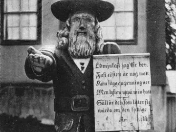 Rosenbom, a wooden statue of a bearded man in a broad-brimmed hat and coat, holding a sign and extending one hand.