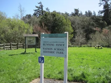 Informational sign for Running Fence in a grassy clearing with a split-rail fence and trees behind it.