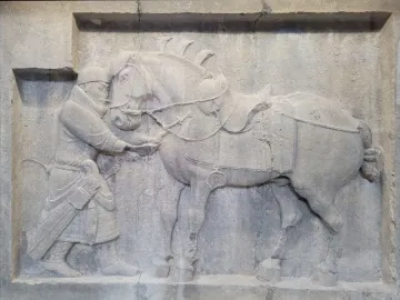 Limestone relief from the Six Steeds of Zhao Mausoleum showing a uniformed figure beside a saddled horse.