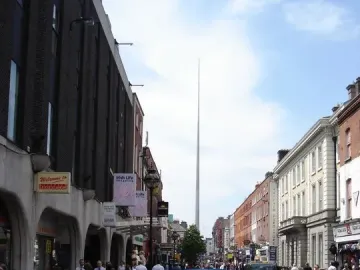 The Spire rises above distant buildings along a busy street with pedestrians and parked cars.