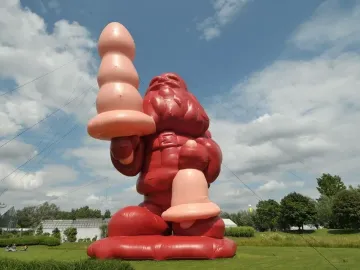 A massive inflatable Santa Claus stands on a grassy lawn holding a large pink object, with another pink object near its left leg.