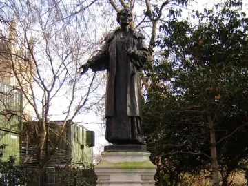 Pankhurst Memorial statue on a tiered stone pedestal in Victoria Tower Gardens, with trees and a low wall behind it.