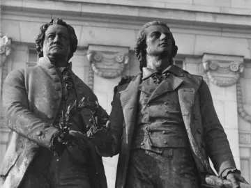 Goethe and Schiller Monument in Weimar, showing two bronze figures in period clothing, one holding a laurel wreath and the other resting an arm on his companion\'s shoulder.