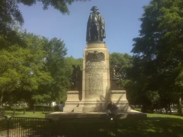 Major General Friedrich Wilhelm von Steuben statue on a tall stone pedestal in a grassy park with trees, with a person sitting at the base behind a low barrier.
