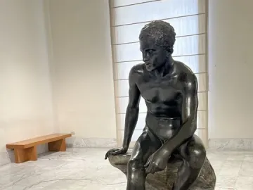 Seated Hermes bronze statue of a nude male figure on a rock, looking downward.