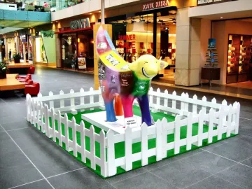 Superlambanana sculpture on artificial grass inside a shopping mall, surrounded by a white picket fence.
