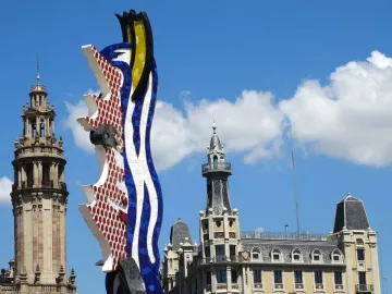 The Face of Barcelona mosaic sculpture rises in front of ornate waterfront buildings under a blue sky.
