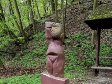 Venus of Petrkovice stone statue on a base beside a gabion wall on a wooded hillside.