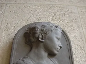 Stone bust of a child in profile, mounted on a light textured wall within an arched frame.