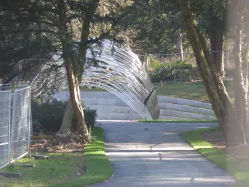 Infinite Wave sculpture spanning a paved walkway in Cannon Hill Park, framed by trees and a fence.
