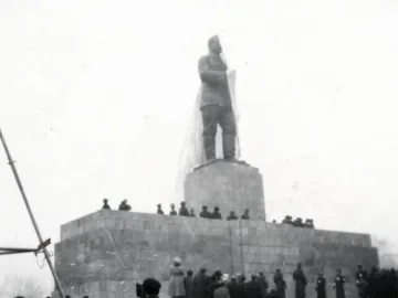 A veiled Stalin monument stands on a tall stone pedestal as a large crowd gathers around it.