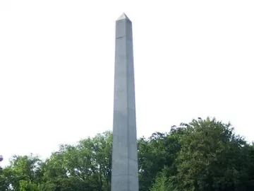 The Needle obelisk stands beside a paved road with a low fence around its base, with trees and a white building in the background.