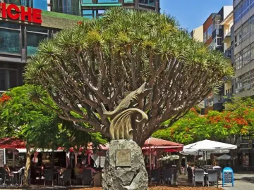 The Chicharro Sculpture, a dark metal fish on a stone pedestal, in front of a large tree and flower bed.