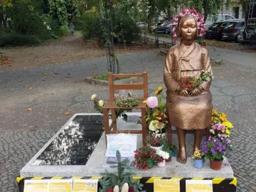 Peace Statue showing a bronze girl seated barefoot on a chair, wearing a flower wreath and holding a small bouquet, surrounded by potted flowers.