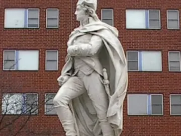 George Washington statue in historical attire standing on a stone pedestal with one foot on a stylized boat.