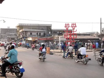 Njé Mo Yé red iron sculpture rising above a busy street with people, motorbikes, and buildings nearby.