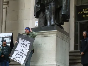 A large George Washington statue stands on a stone pedestal while people gather nearby, including one holding a sign.
