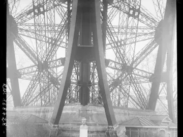 Bust of Gustave Eiffel on a tall pedestal, with floral wreaths and a crowd gathered at the base of the Eiffel Tower.