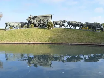 The Monument to the Cart, a bronze sculpture of a covered cart pulled by oxen with a rider on horseback nearby, reflected in a pond.