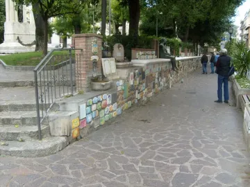 The Muretto of Alassio stone wall decorated with colorful ceramic tiles, a bronze figure perched on top, and a small crest pillar near steps.