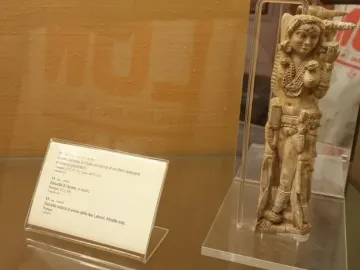 Ivory Lakshmi statuette with elaborate jewelry, headdress, and carved figures at the sides, displayed upright in a clear case.
