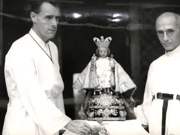 Santo Niño de Cebú statue on an ornate pedestal, flanked by two men in clerical attire.