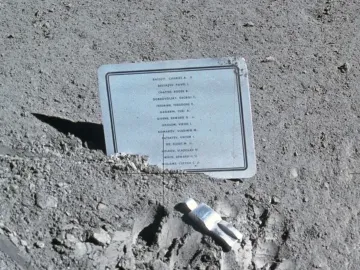 A small fallen astronaut figure lies on the lunar surface beside a memorial plaque.
