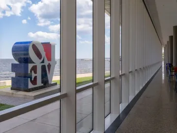 The Love sculpture is seen outdoors through large museum windows beside the water under a cloudy sky.