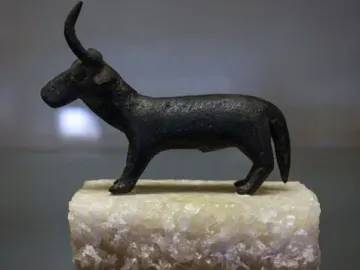 A dark Nuragic bronze statuette of a horned animal on a light rectangular base, shown with a reflected image on a glossy surface.