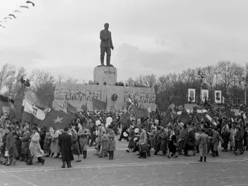 Crowd parades past the Stalin Monument, with flags, balloons, and portrait banners around the towering statue.