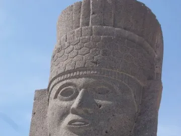 Close-up of an Atlantean figure at Tula with a carved face and patterned headdress against a blue sky.