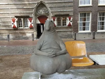 Homunculus Loxodontus sculpture seated on a metal bench outside, with a brick building behind it.