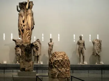 Dancers of Delphi sculpture with three draped female figures on a carved column base in a museum hall.