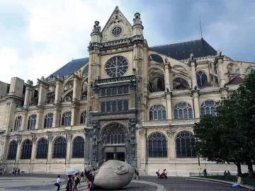 Écoute, a pale stone head sculpture with a hand cupped to its ear, stands in a plaza with a church behind it.