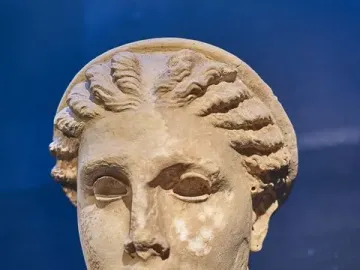 Weathered stone head of Lycosoura Artemis with braided hair on a small pedestal.