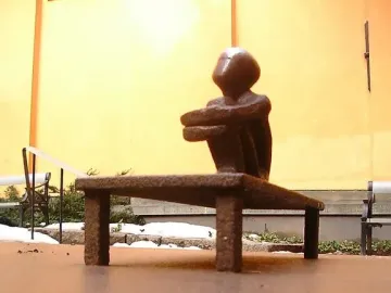 Järnpojke, a small dark seated sculpture on a low bench beside a building wall with patches of snow and greenery.