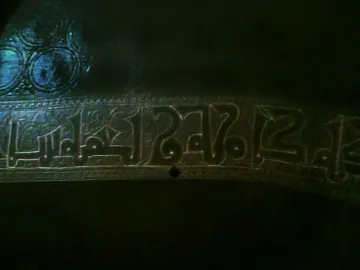 Close-up of the Pisa Griffin showing a band of raised Kufic calligraphy and circular motifs on the bronze surface.