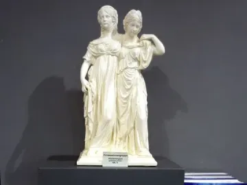 Small white Princesses Monument statuette of two women in classical dresses, displayed on a pedestal among books and souvenirs.