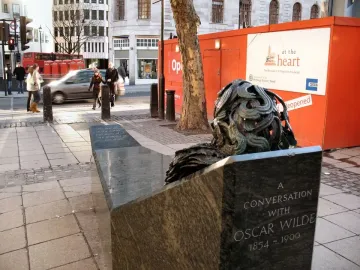A Conversation with Oscar Wilde bench sculpture with an abstract bronze-toned bust and swirling hair on a city sidewalk.