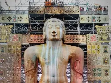 Gommateshwara Bahubali statue with ritual streaks, framed by a decorated scaffolding structure and a smiling man in orange robes in the foreground.