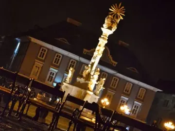 Holy Trinity Monument illuminated at night above wet cobblestones and park benches