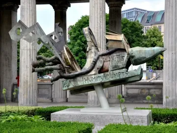 Humpty Dumpty Machine bronze sculpture with a green and brown patina, shown outdoors on a plinth beside classical columns.